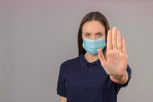 Young woman wearing blue polo shirt in protective medical mask showing hand stop gesture with serious face isolated on light grey background with copy space