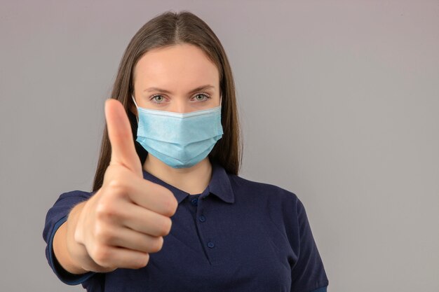 明るい灰色の背景の上に立って肯定的な表現を親指を示す防護医療マスクで青いポロシャツを着た若い女性