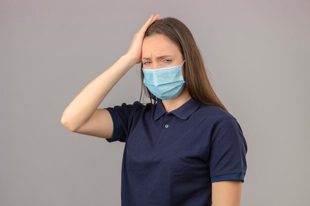 Young woman wearing blue polo shirt in protective medical mask touching head feeling headache isolated on light grey background