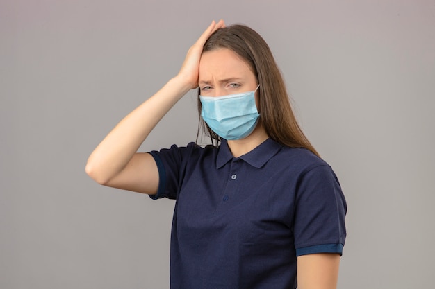 Молодая женщина в синей рубашке поло в защитной медицинской маске, касаясь головы, чувствуя головную боль, изолированные на светло-сером фоне