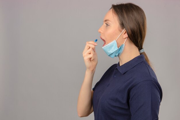 Молодая женщина в синей рубашке поло в защитной медицинской маске с открытым ртом, принимая таблетки на светло-сером фоне