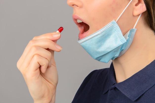 Young woman wearing blue polo shirt in protective medical mask with open mouth taking a pill on light grey background