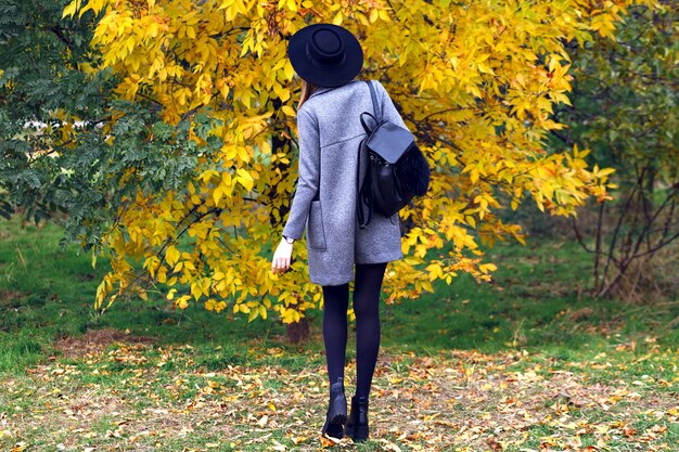 Young woman wearing casual elegant street style outfit, hat and coat walking in autumn day city park, posing back.