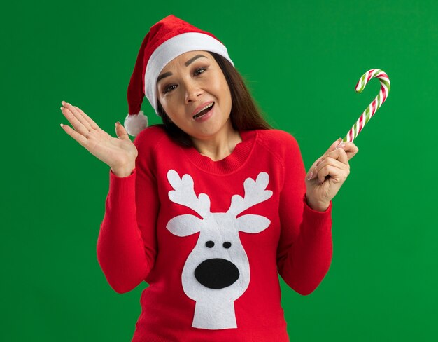 Free photo young woman wearing christmas santa hat and red sweater holding candy cane  looking at camera happy and cheerful standing over green background
