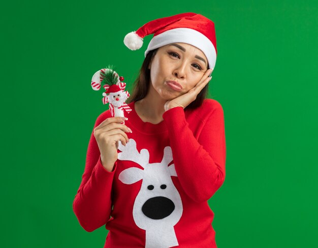 Young woman wearing christmas santa hat and red sweater holding christmas candy cane looking at camera with sad expression  standing over green background