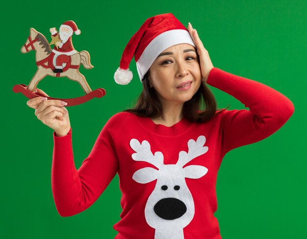 クリスマスのサンタの帽子と赤いセーターを着て、緑の背景の上に立っている彼女の頭の上の手と混乱しているカメラを見てクリスマスのおもちゃを保持している若い女性