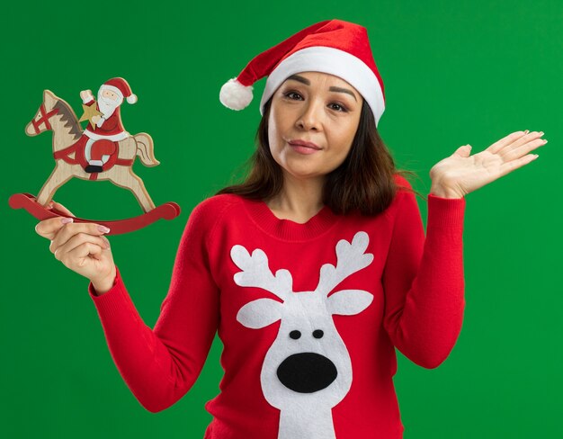 緑の背景の上に立っている顔に笑顔でカメラを見てクリスマスのおもちゃを保持しているクリスマスサンタ帽子と赤いセーターを着ている若い女性