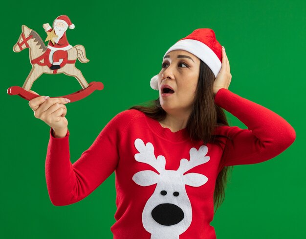 Young woman wearing christmas santa hat and red sweater holding christmas toy  looking at it surprised standing over green background