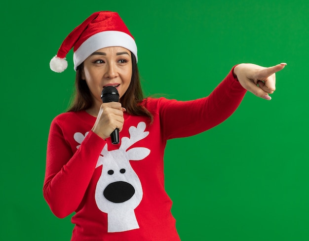 Giovane donna che indossa il natale santa hat e maglione rosso parlando al microfono guardando a parte con la faccia felice che punta con indice figner al lato in piedi su sfondo verde