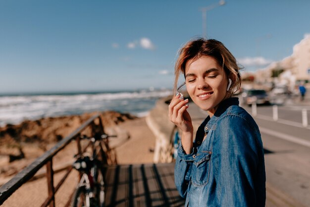 ビーチでポーズをとって電話でエアポッドとデニムシャツを着ている若い女性