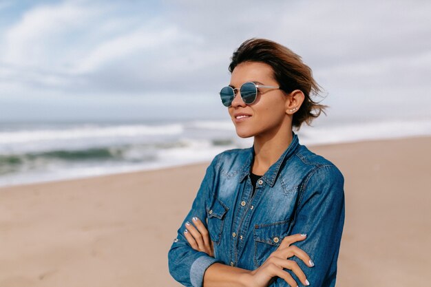 Молодая женщина в джинсовой рубашке с солнцезащитными очками позирует на пляже