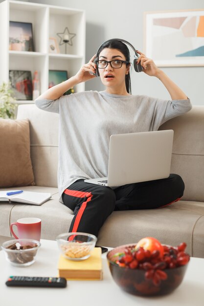 リビングルームのコーヒーテーブルの後ろのソファに座ってヘッドフォンを保持し、ラップトップを使用して眼鏡をかけている若い女性