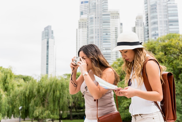 Шляпа молодой женщины нося читая карту и ее подруга фотографируя от камеры