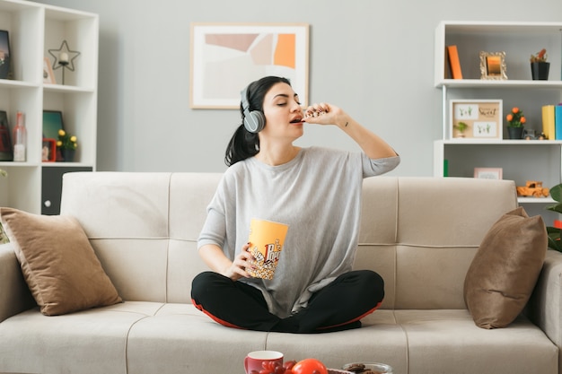 Young woman wearing headphones eat popcorn sitting on sofa behind coffee table in living room