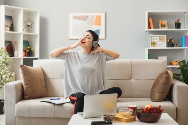 リビングルームのコーヒーテーブルの後ろのソファに座っているヘッドフォンを身に着けている若い女性