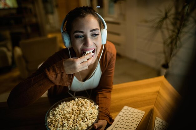 ポップコーンを食べて、自宅のコンピューターでインターネット上で何かを見ながらヘッドフォンを身に着けている若い女性
