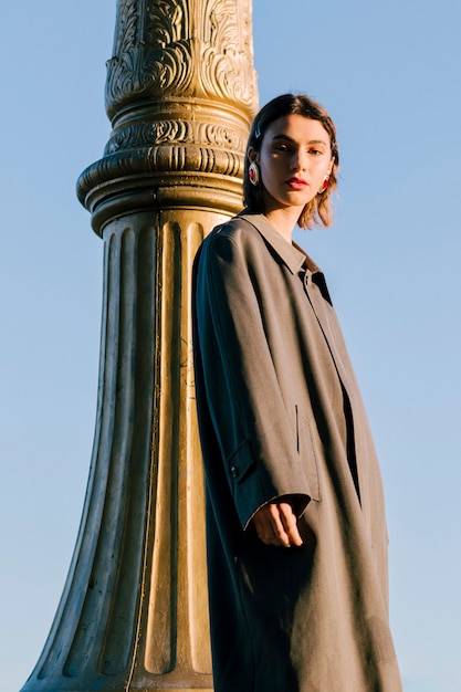 Free photo young woman wearing long coat standing near the pillar against blue sky