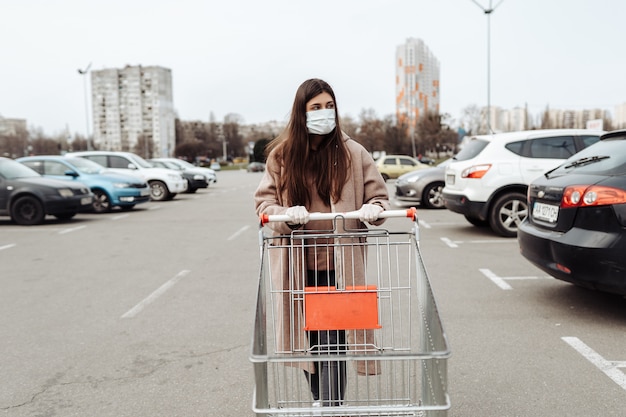 Молодая женщина носить защитную маску против коронавируса 2019-нКоВ, толкая корзину.