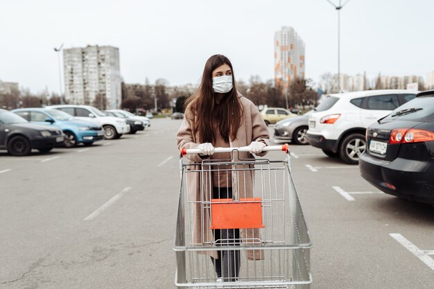 쇼핑 카트를 밀고 코로나 바이러스 2019-nCoV에 대 한 보호 얼굴 마스크를 착용하는 젊은 여자.
