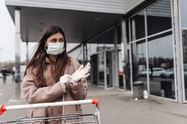 Молодая женщина носить защитную маску против коронавируса 2019-нКоВ, толкая корзину.