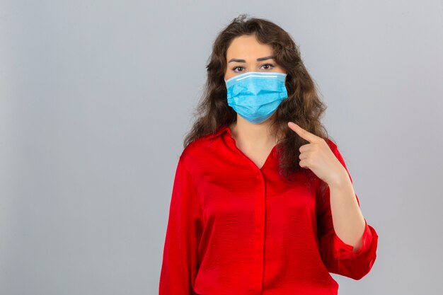 Молодая женщина в красной блузке в медицинской защитной маске смотрит в камеру и указывает пальцем на маску на лице на изолированном белом фоне
