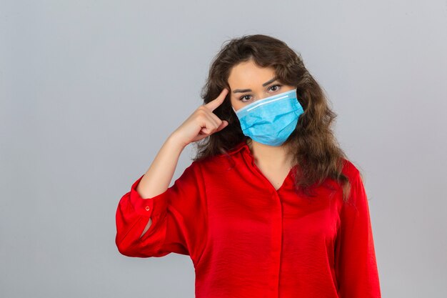 Young woman wearing red blouse in medical protective mask looking at camera pointing with finger to her head over isolated white background