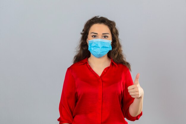 Young woman wearing red blouse in medical protective mask looking at camera with smile showing thumb up over isolated white background