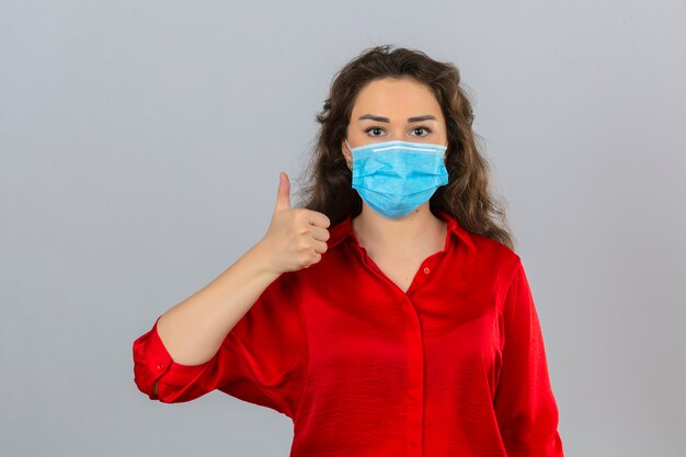 Young woman wearing red blouse in medical protective mask looking at camera with smile showing thumb up over isolated white background