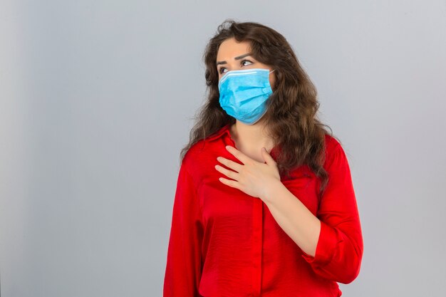 Молодая женщина в красной блузке в медицинской защитной маске выглядит нездоровой и больной с рукой на груди на изолированном белом фоне