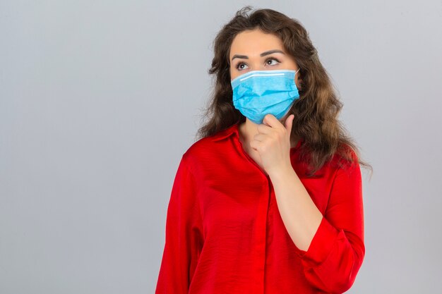 Молодая женщина в красной блузке в медицинской защитной маске, стоя с рукой на подбородке, думает, что сомневается в изолированном белом фоне