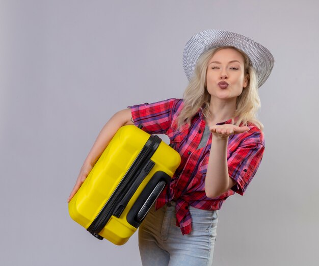 Young woman wearing red shirt in hat holding suitcase showing kiss gesture on isolated white wall