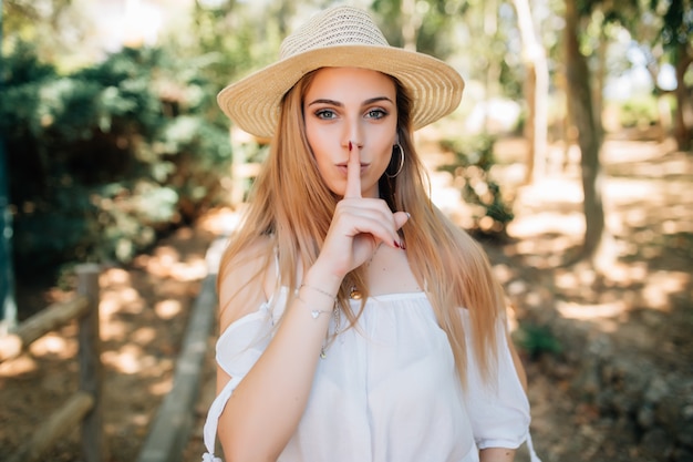 Young woman wearing summer hat with index finger on lips, ask to be quiet. Silence and secret concept