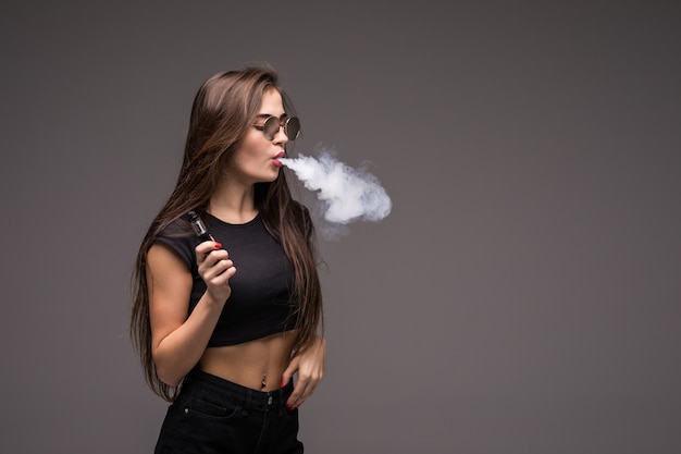 Young woman wearing in sunglasses smoking vape on gray wall.