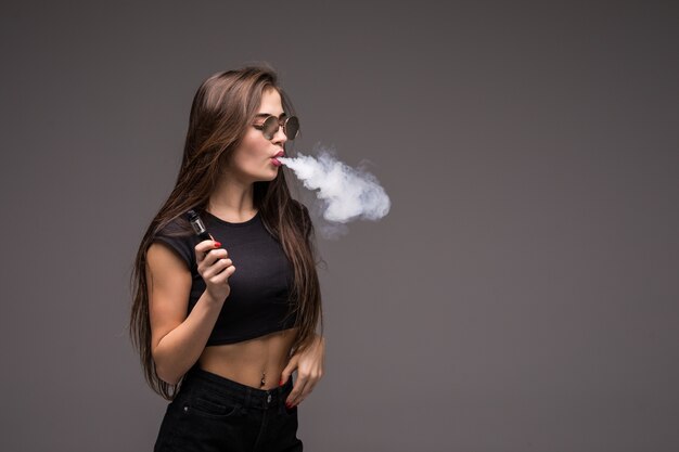 young woman wearing in sunglasses smoking vape on gray wall.