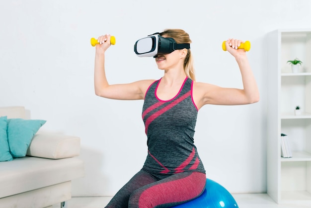 Молодая женщина, носить гарнитуру виртуальной реальности, сидя на фитнес-мяч, работающ с желтыми гантелями