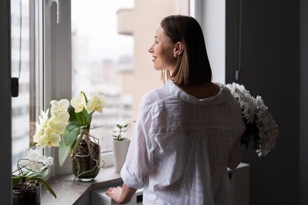 Молодая женщина в белой повседневной одежде смотрит в окно с букетом белых цветов в ожидании весны или лета