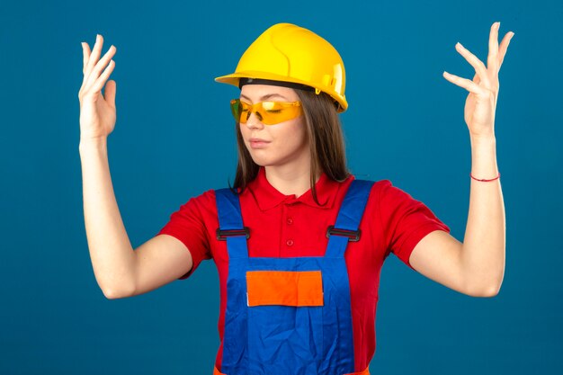 Молодая женщина в желтых очках в строительной форме и желтый защитный шлем, стоя с закрытыми глазами и поднятыми руками на синем фоне