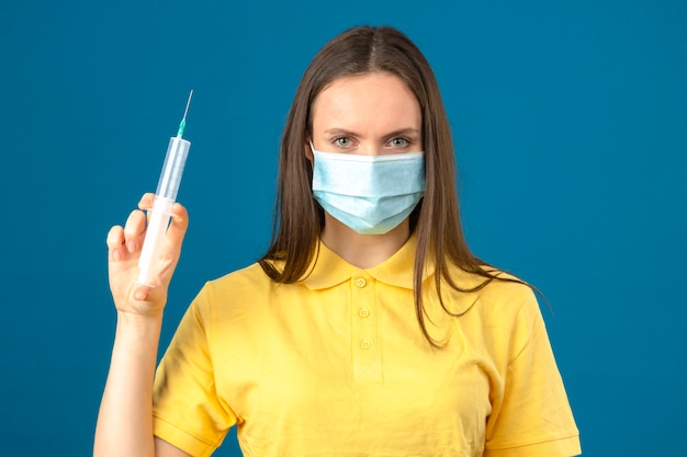 孤立した青い背景に深刻な顔立ちでカメラを見て注射器を保持している防護医療マスクで黄色のポロシャツを着た若い女性
