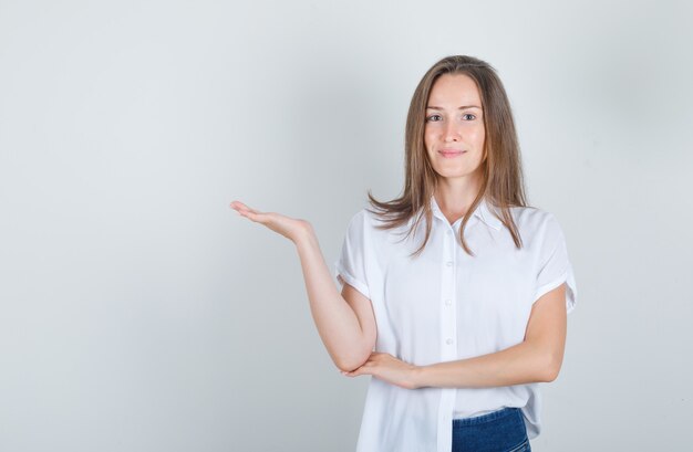 白いTシャツ、ジーンズで何かを歓迎または見せて、陽気に見える若い女性