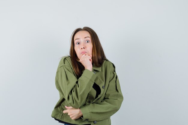 Young woman while holding her fist on jaw in green jacket and looking attracted , front view.