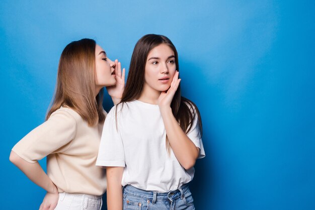 Young woman whispers to her mate bad news isolated on blue wall