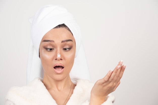 Young woman in a white bathrobe applying a cream on her nose .