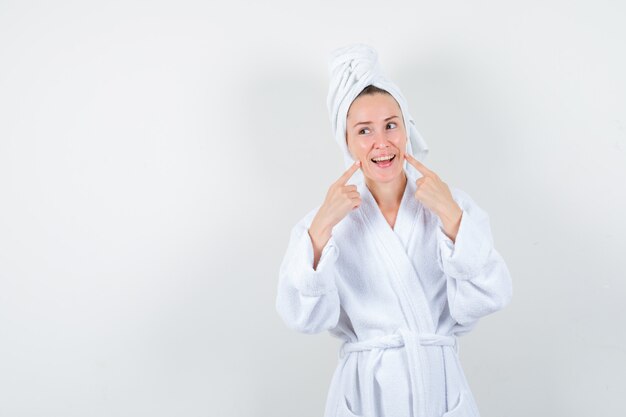 Молодая женщина в белом халате, полотенце держит пальцы на щеках и выглядит весело, вид спереди.