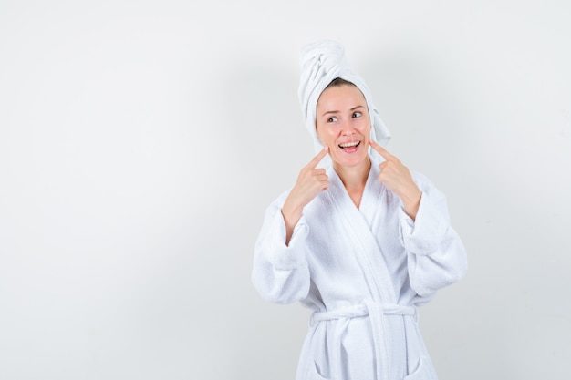 Young woman in white bathrobe, towel keeping fingers on cheeks and looking jolly , front view.