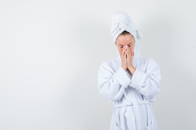 Молодая женщина в белом халате, полотенце, держа руки в молящемся жесте и выглядящем обнадеживающим, вид спереди.