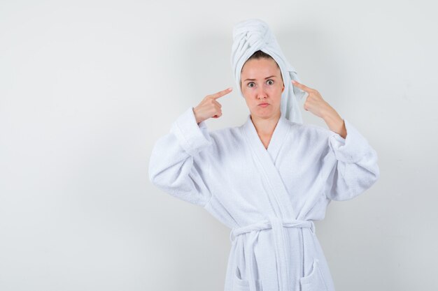 Young woman in white bathrobe, towel pointing at her head and looking puzzled , front view.