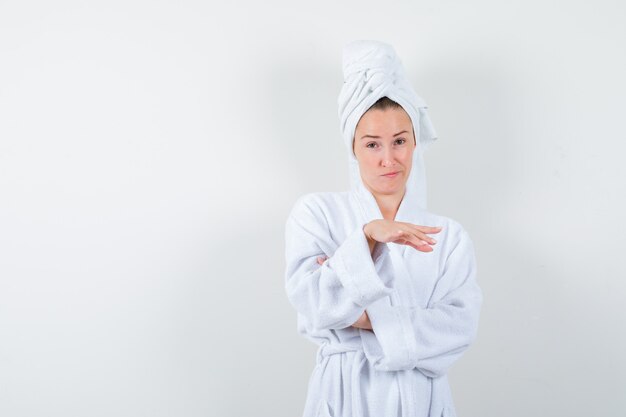 Молодая женщина в белом халате, полотенце позирует, прикрывая что-то ладонью и выглядя сомнительно, вид спереди.