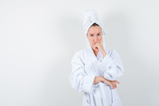 Young woman in white bathrobe, towel propping chin on hand and looking sensible , front view.