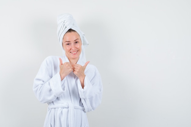 Young woman in white bathrobe, towel showing double thumbs up and looking cheerful , front view.