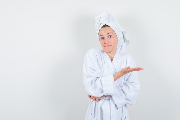 Young woman in white bathrobe, towel spreading palm to show something and looking puzzled , front view.